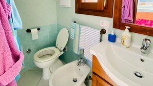 Un baño con inodoro, bidé y lavamanos. Hay un perchero con toallas, un espejo y una ventana con vista a la playa.