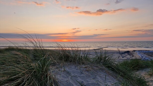 Une plage avec des dunes de sable et de l’herbe au premier plan, une mer calme et un coucher de soleil en arrière-plan.