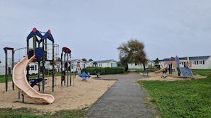 Ein Spielplatz mit verschiedenen Spielgeräten, darunter Rutschen und Klettergerüsten, umgeben von einem gepflasterten Weg und einer Rasenfläche.