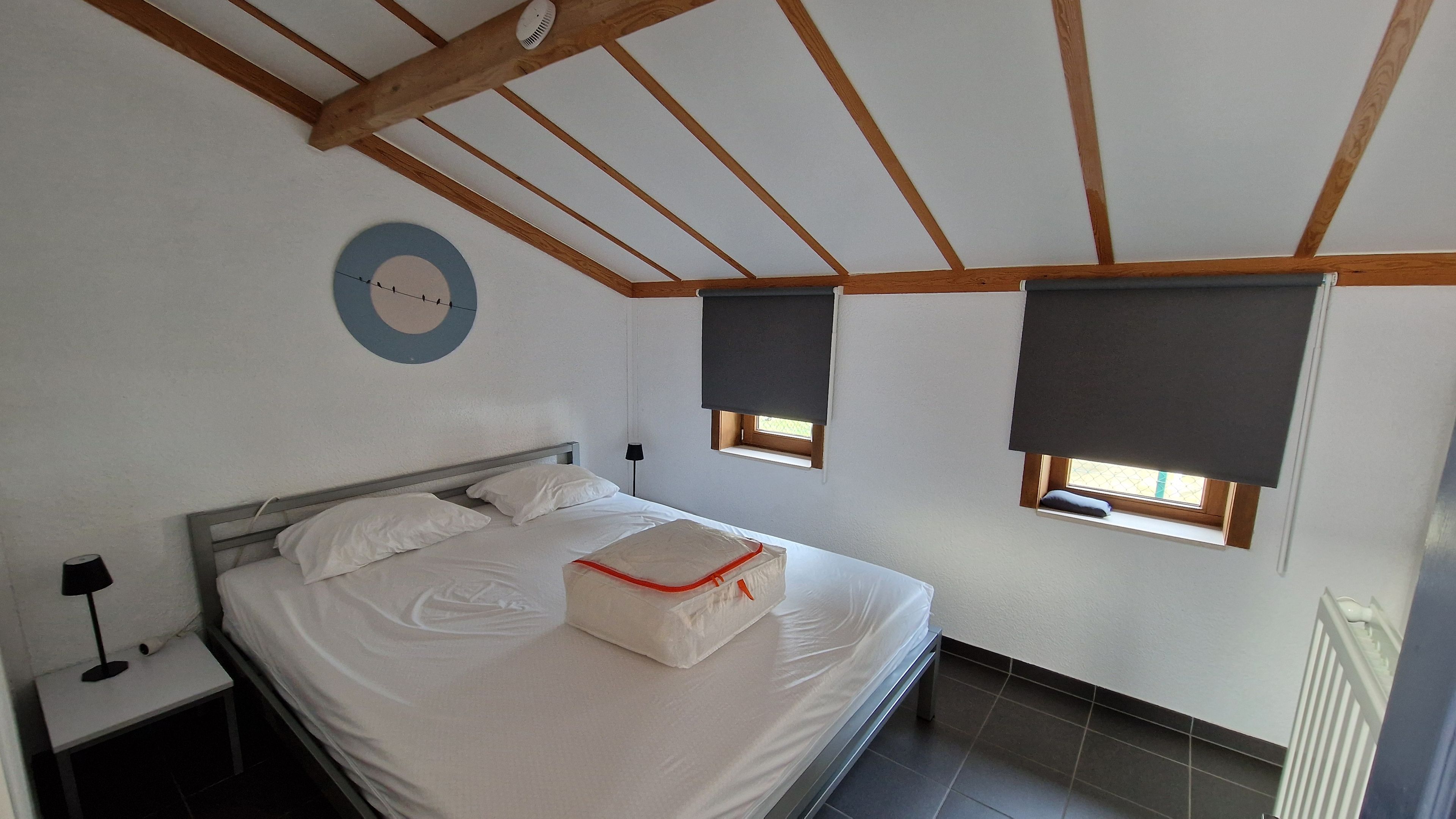 A bedroom with a bed, a bedside table, a lamp, a circular wall decoration, and two windows with blinds.
