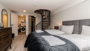 A bedroom with a large bed, a dresser, a mirror, a spiral staircase, and a washing machine in the background.