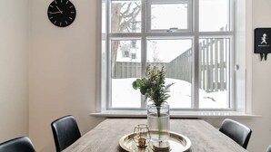 Un comedor con una mesa de madera, sillas, una bandeja con vasos, un reloj y una ventana que da a un patio nevado.