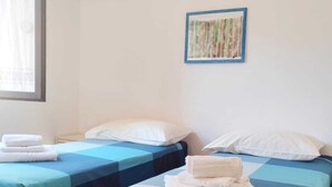A room with two beds, each with a blue and white checkered bedspread, white pillows, and folded towels on the beds. There is a window with curtains, an air conditioning unit on the wall, and a framed picture above the beds. - Arianna House (Borgo Rocce Rosse Arbatax) (Arbatax)