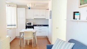 A compact kitchen with white cabinets, a dining table, and chairs. There is a blue sofa in the living area. - Arianna House (Borgo Rocce Rosse Arbatax) (Arbatax)