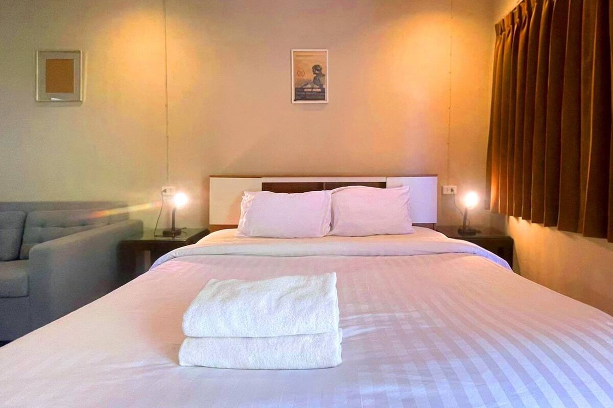 A neatly made bed with pillows and a folded towel, a bedside lamp, and a framed picture on the wall.