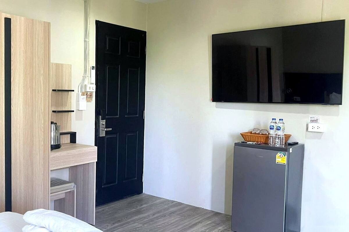A hotel room with a bed, a desk with a kettle, a refrigerator, and a black door.