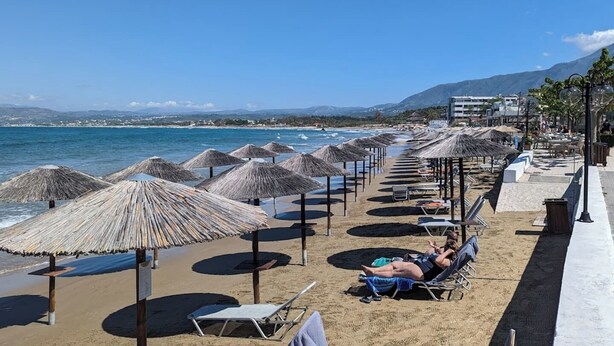 Una platja amb files de gandules i sombrerets de palla, un cel serè i una vista al mar i muntanyes llunyanes.