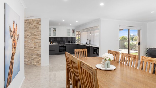 A modern kitchen with a dining area, featuring a stone accent wall, a wooden dining table, and a view of the backyard through glass doors. - Heinlen on North Terrace  (Mount gambier)