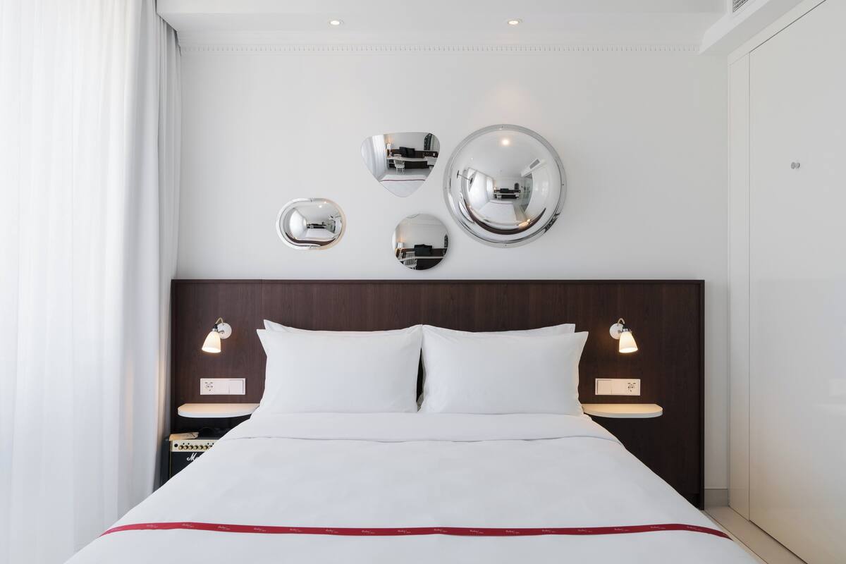 A hotel room with a neatly made bed, a wooden headboard, bedside tables with lamps, and three wall mirrors.