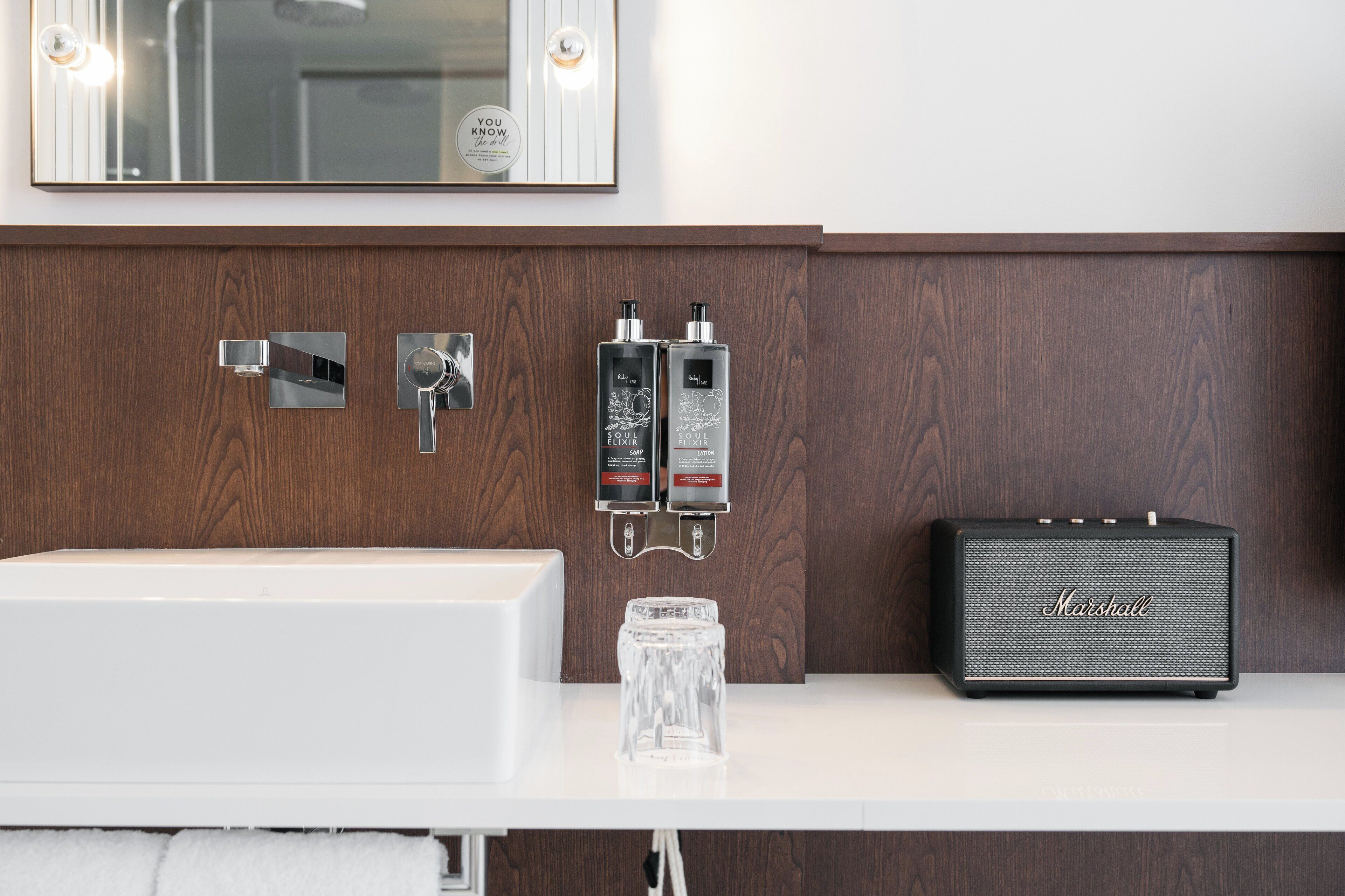 Un baño con tocador de madera, un lavabo blanco, dos botellas de jabón para manos, un vaso y un amplificador Marshall.