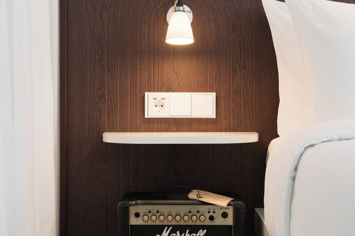 A bedroom with a wooden headboard, a bedside lamp, a Marshall amplifier, and a bed with white bedding.