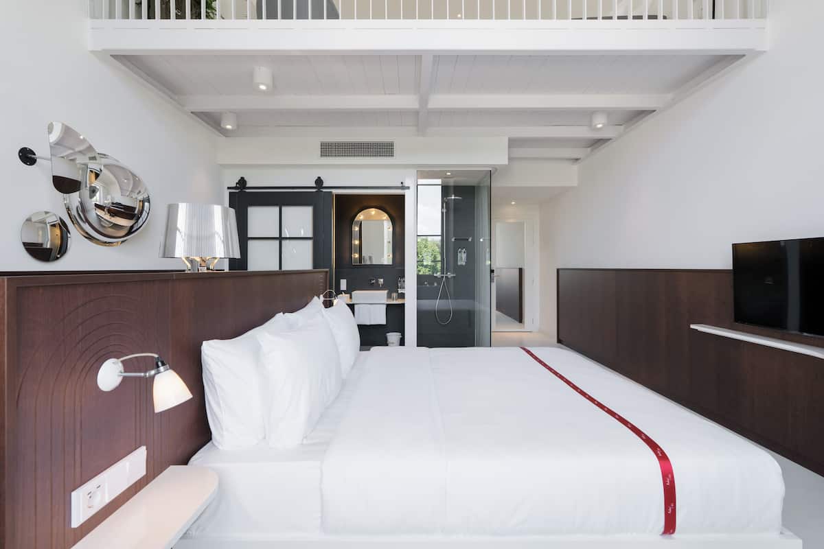 A modern hotel room with a large bed, a TV, a bathroom with a glass door, and a balcony.