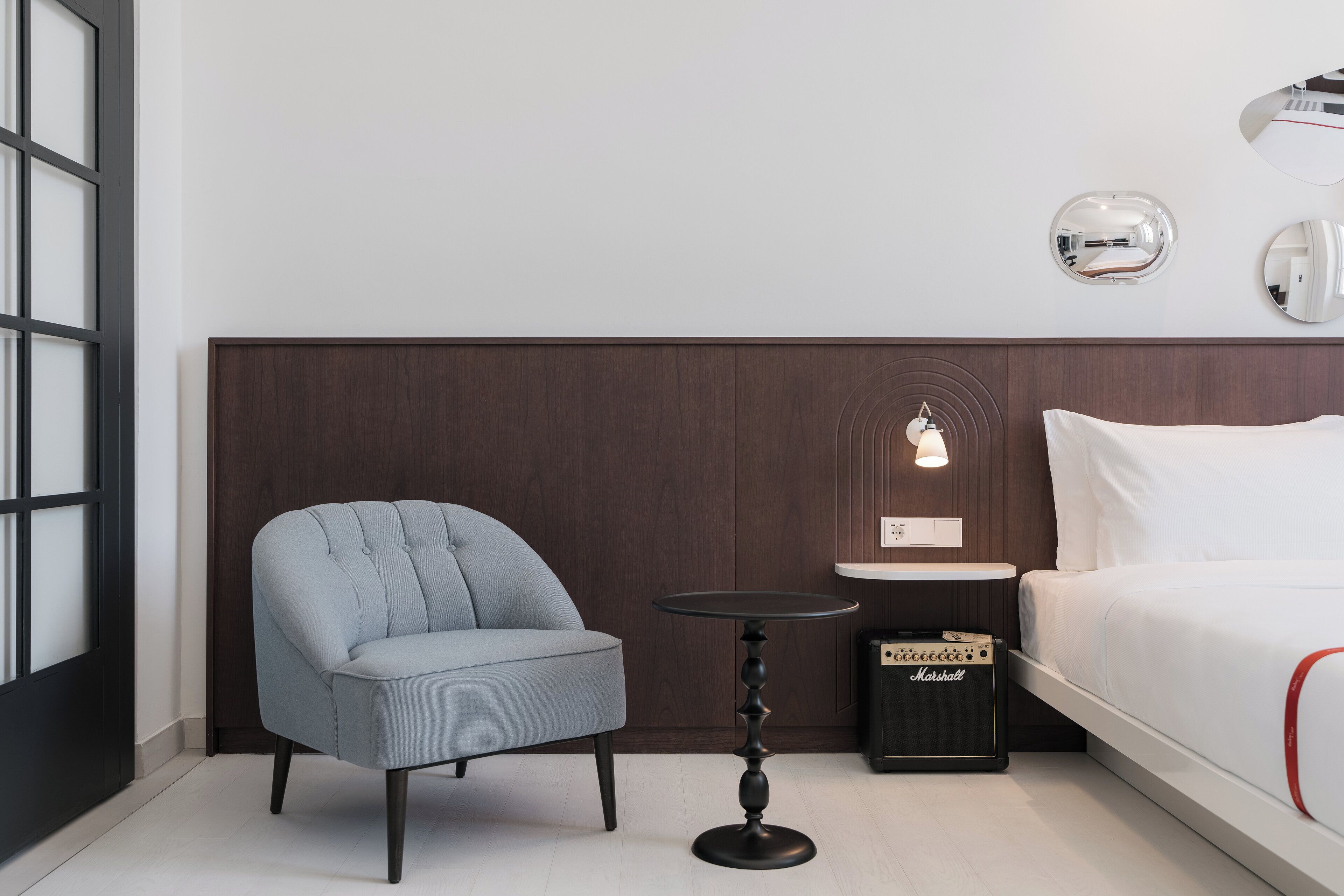 Een moderne hotelkamer met een grijs gestoffeerde fauteuil, een klein rond tafeltje, een Marshall-versterker en een bed met wit beddengoed.