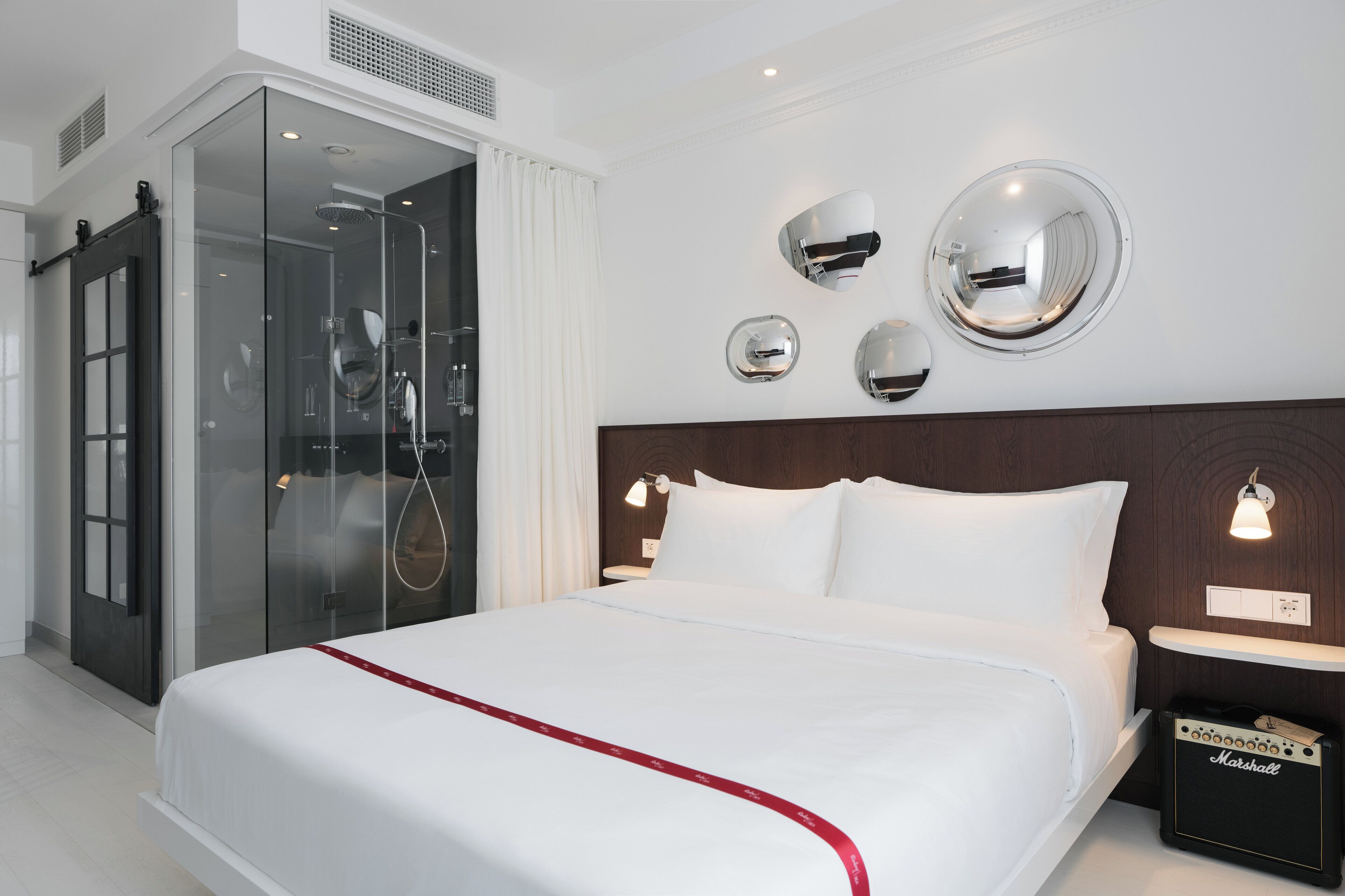 A modern hotel room with a large bed, a glass shower enclosure, and a Marshall amplifier on the nightstand.