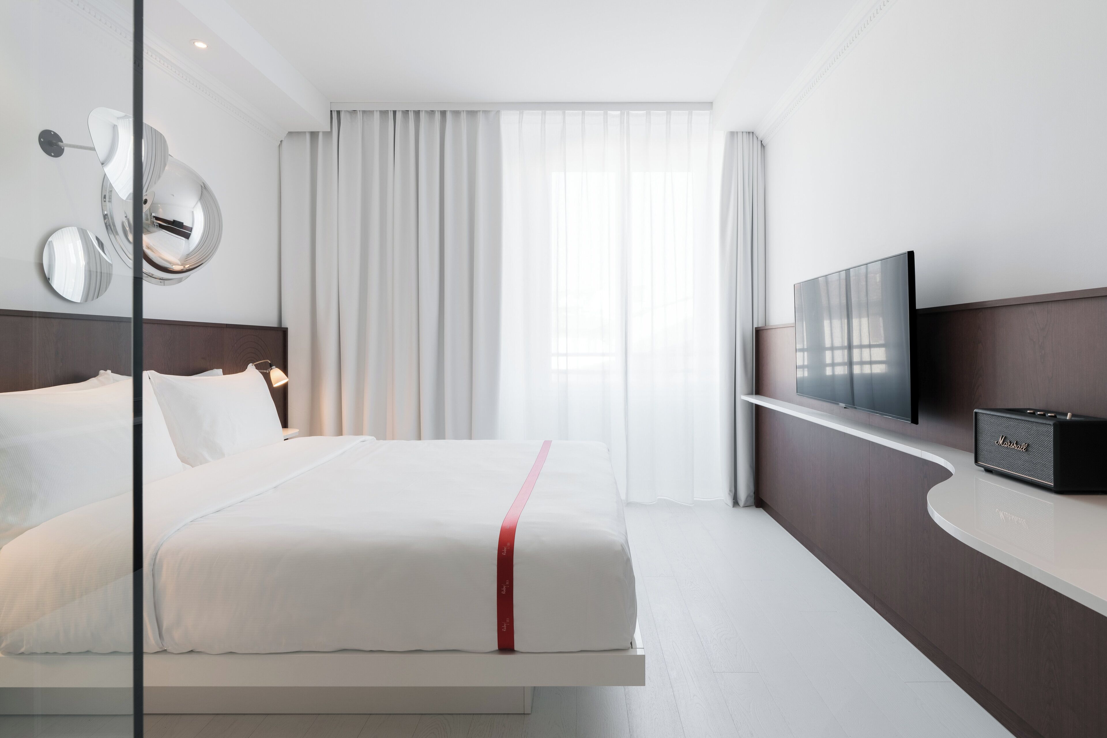 A modern hotel room with a bed, a flat-screen TV, a bedside table, and a large window with curtains.
