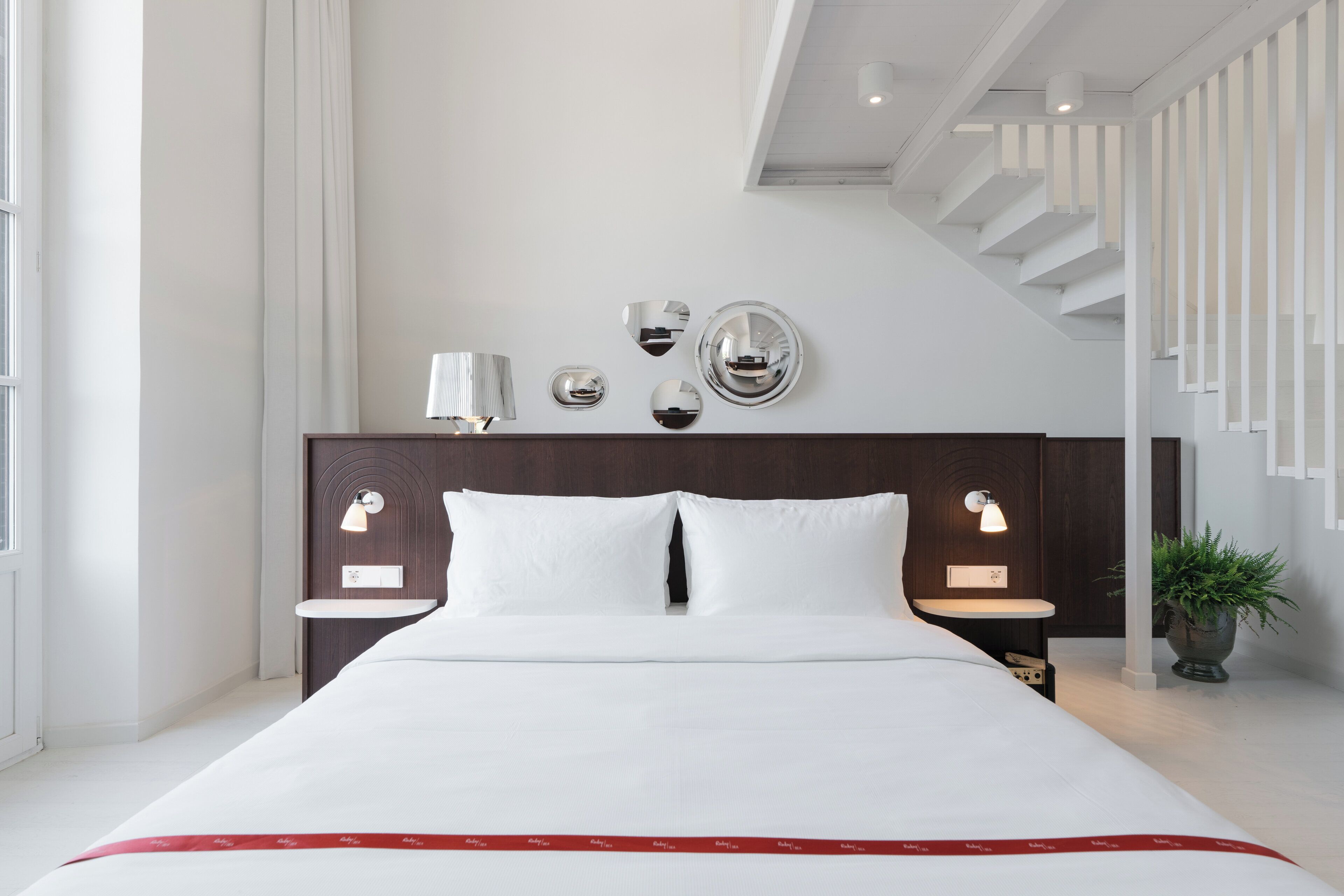 Ein modernes Hotelzimmer mit einem großen Bett, einem Kopfteil mit integrierter Beleuchtung, einem kleinen Nachttisch und einer Topfpflanze.