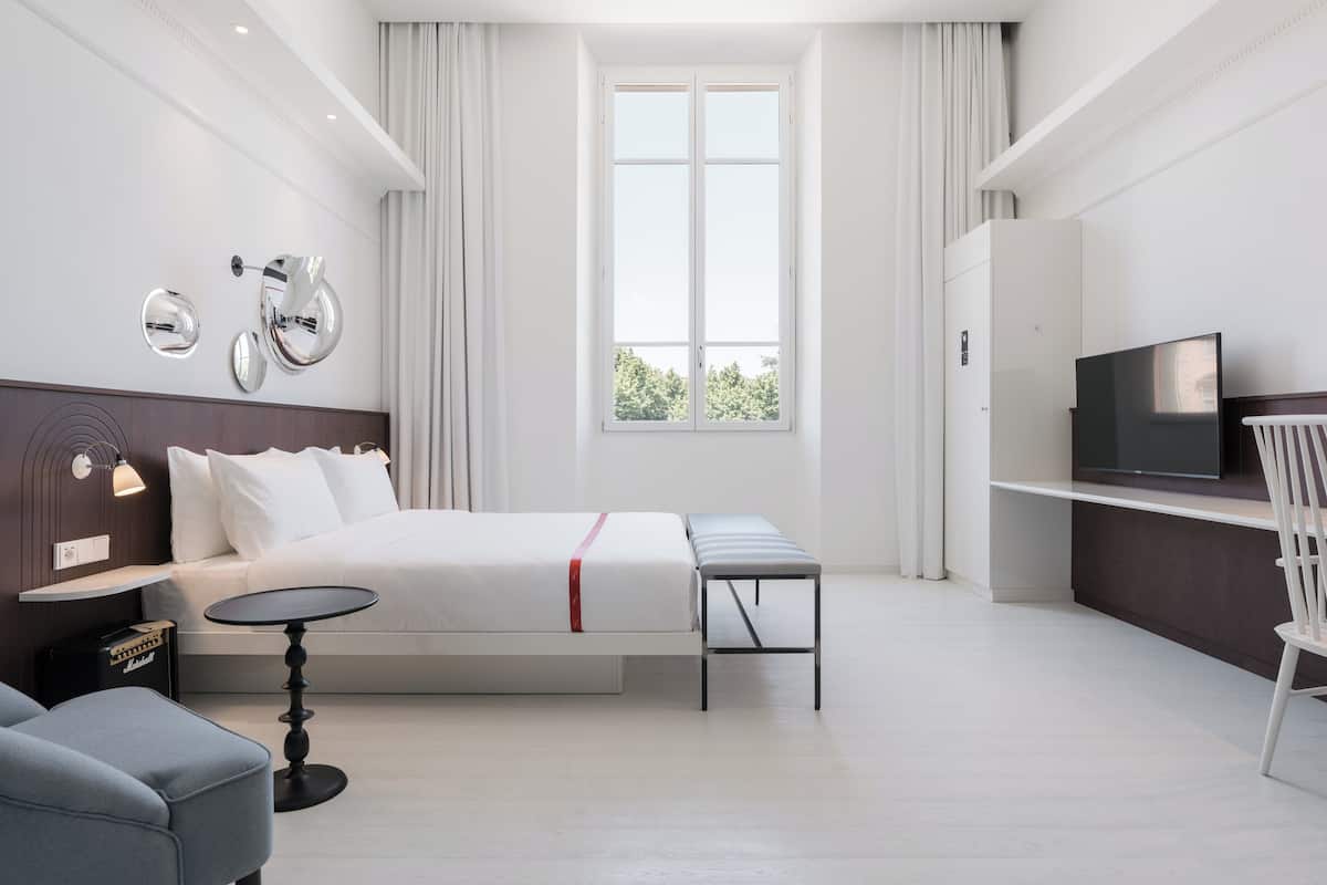 A modern hotel room with a bed, a desk, a television, and a window with a view of greenery.