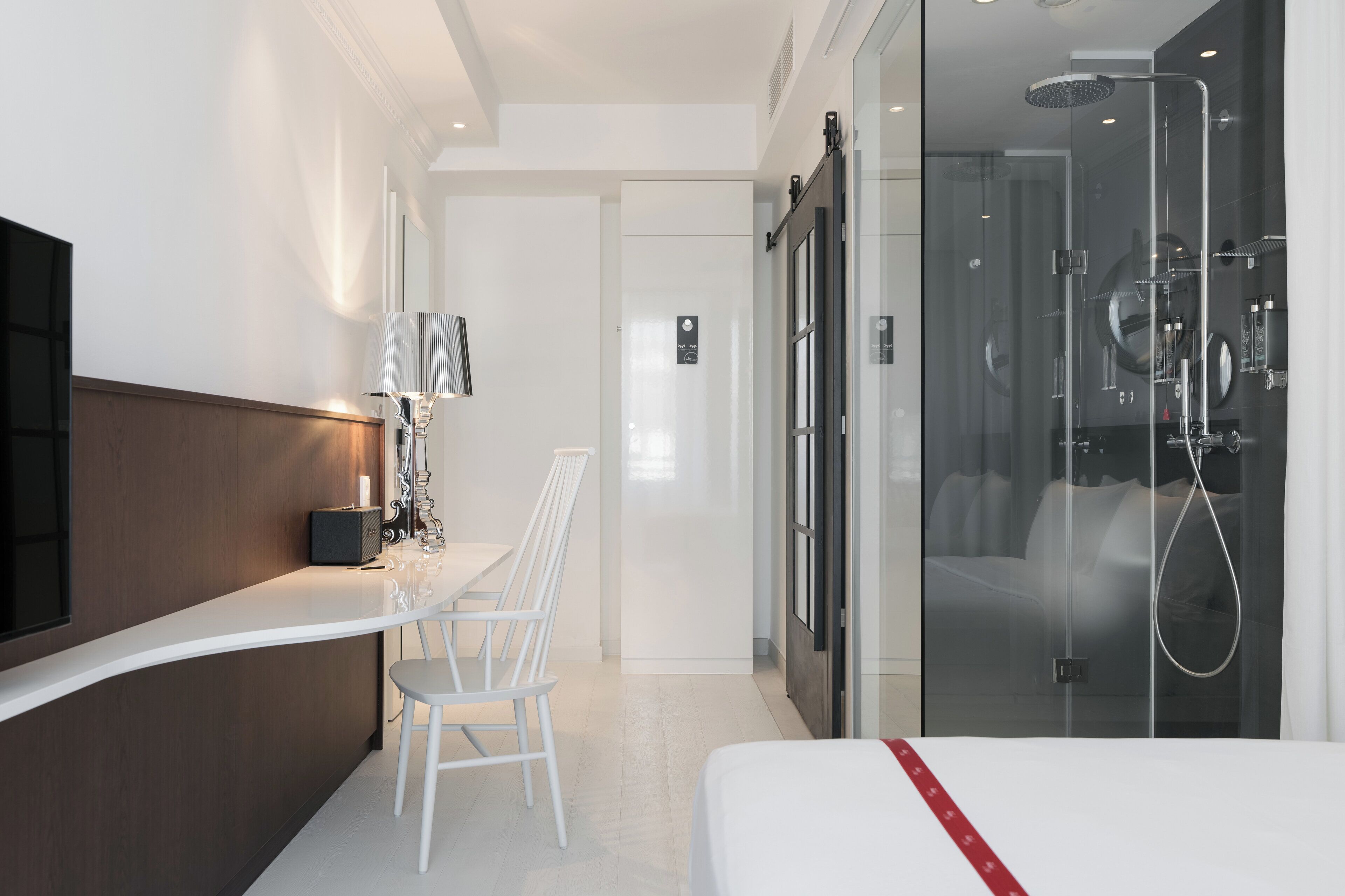 A modern hotel room with a curved white counter, a single white chair, a bed with a red-striped bedspread, and a glass-enclosed shower area.
