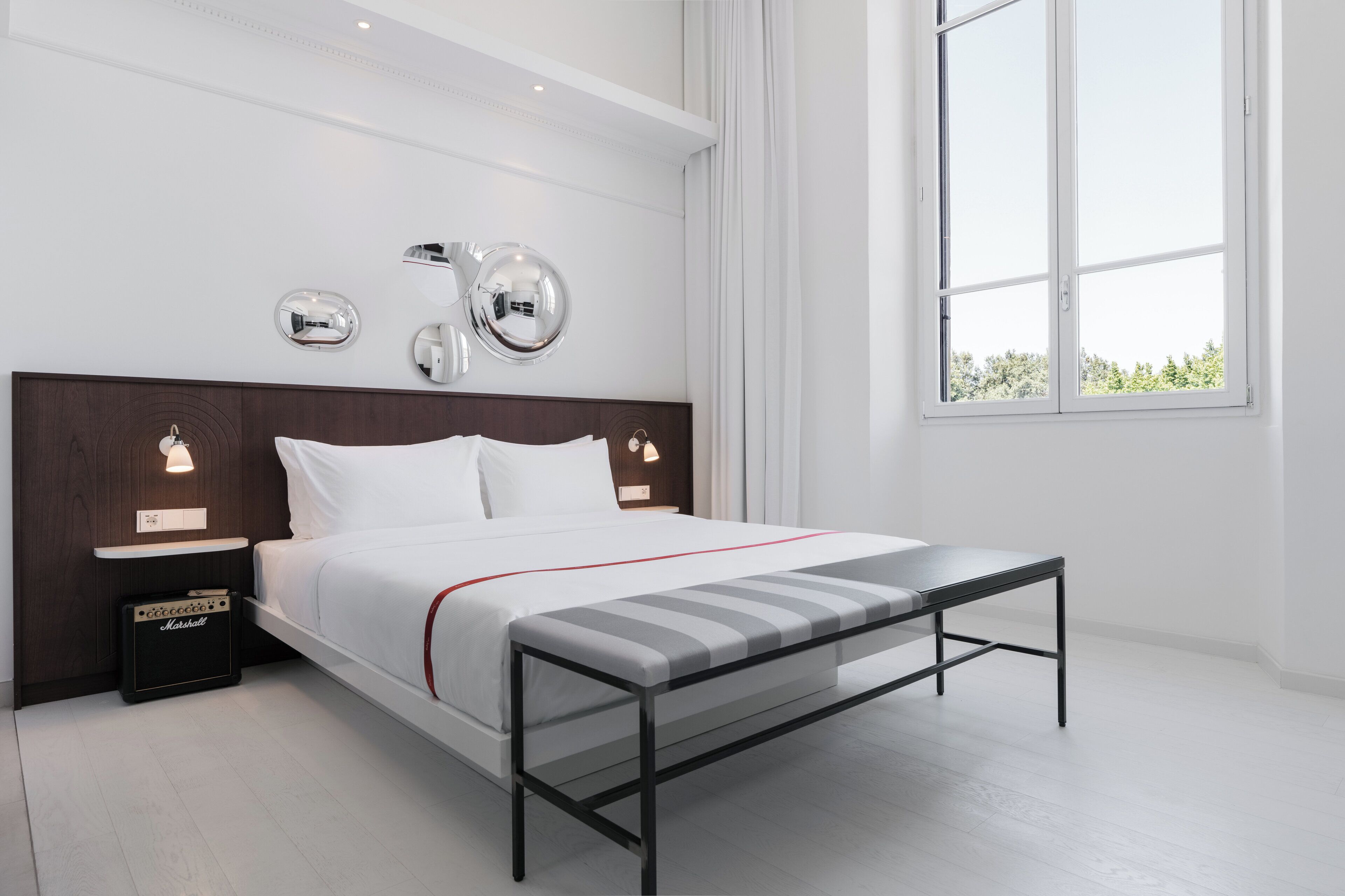 A modern hotel room with a large bed, bedside tables with lamps, a bench, and a large window with a view.