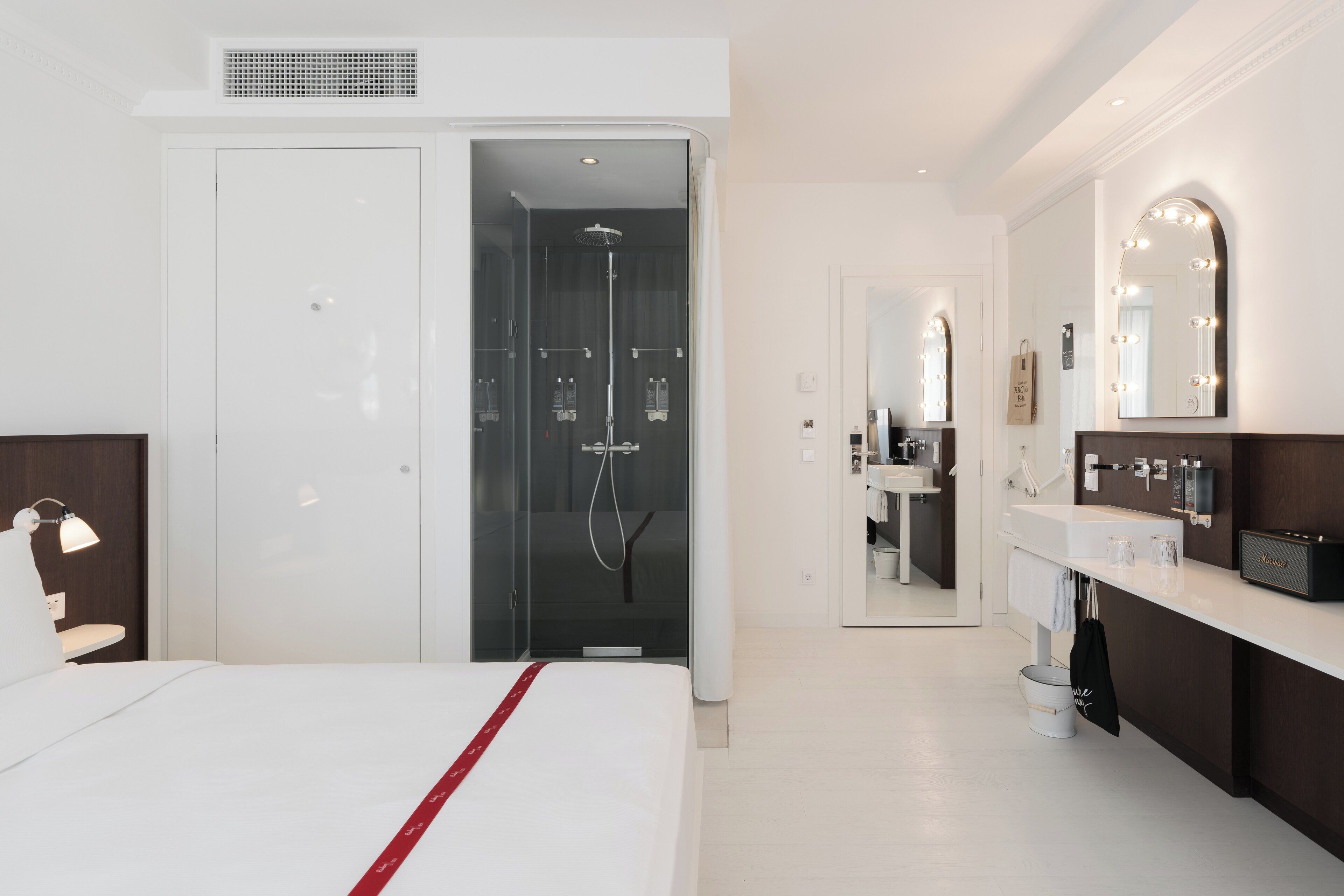 A modern hotel room with a large bed, a separate bathroom with a shower, a vanity with a mirror, and a small seating area with a lamp.