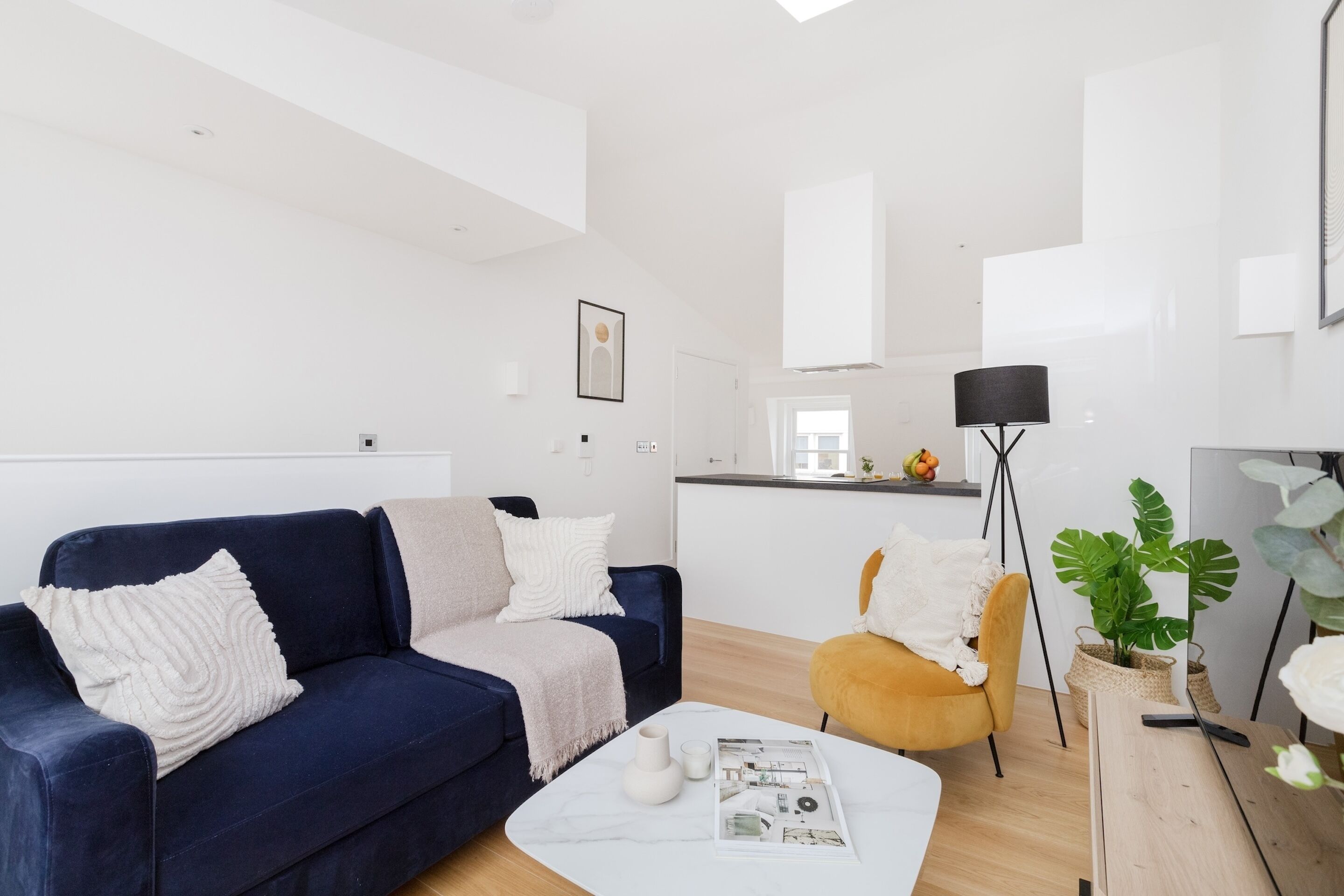 Una sala moderna con un sofá azul oscuro, una mesa de centro blanca y un sillón amarillo. Se observa una zona de cocina al fondo, con una lámpara de pie negra y una planta grande.