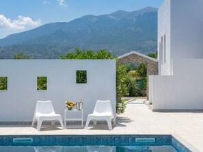 A poolside area with two white chairs, a small table with fruit and a metal container, and a view of mountains in the background. - Lilu Villa 1 With Private Pool and Seaview (Kos)