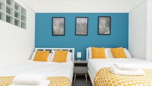 Habitación doble con dos camas, cada una con cubrecama blanco y amarillo estampado, una mesita de noche con lámpara, y tres cuadros enmarcados sobre las camas.