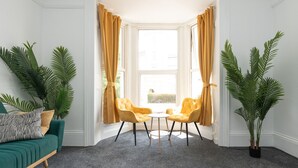 Una habitación con un sofá verde, dos sillas amarillas, una mesita y plantas verdes grandes.