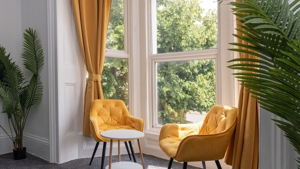 Una habitación con dos sillones amarillos y una pequeña mesa redonda, ventanas amplias con cortinas, y una planta en maceta.