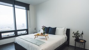 Ein modernes Schlafzimmer mit einem großen Bett, einem Nachttisch mit Lampe, einem Fenster mit Blick auf die Stadt und einer kleinen Topfpflanze.