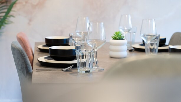 A dining table set with clear glasses, plates, and cutlery, with a small potted plant and a marble wall in the background.