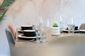 A dining table set with clear glasses, plates, and cutlery, with a small potted plant and a marble wall in the background. - Open Mind Property - 4 Bedroom Lovely Home (Portsmouth)