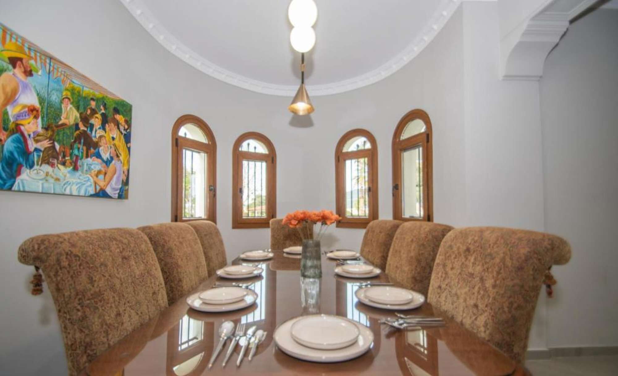 Uma sala de jantar com uma mesa redonda posta para seis pessoas, ladeada por seis cadeiras estofadas. Há um quadro na parede acima da mesa e um vaso com flores sobre a mesa.
