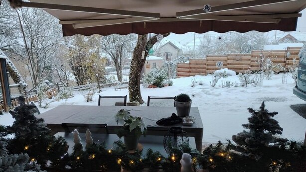 A covered outdoor patio with a table and chairs, surrounded by snow-covered plants and a wooden fence. - Location en Savoie (Arith)