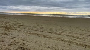 Une vaste plage de sable où l’on peut voir des traces de pneus et des empreintes de pas, qui mènent à l’océan sous un ciel nuageux.