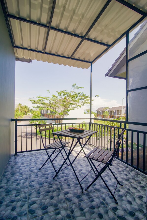 Un balcone coperto con tavolo e sedie, pavimento piastrellato e vista su edifici e alberi.