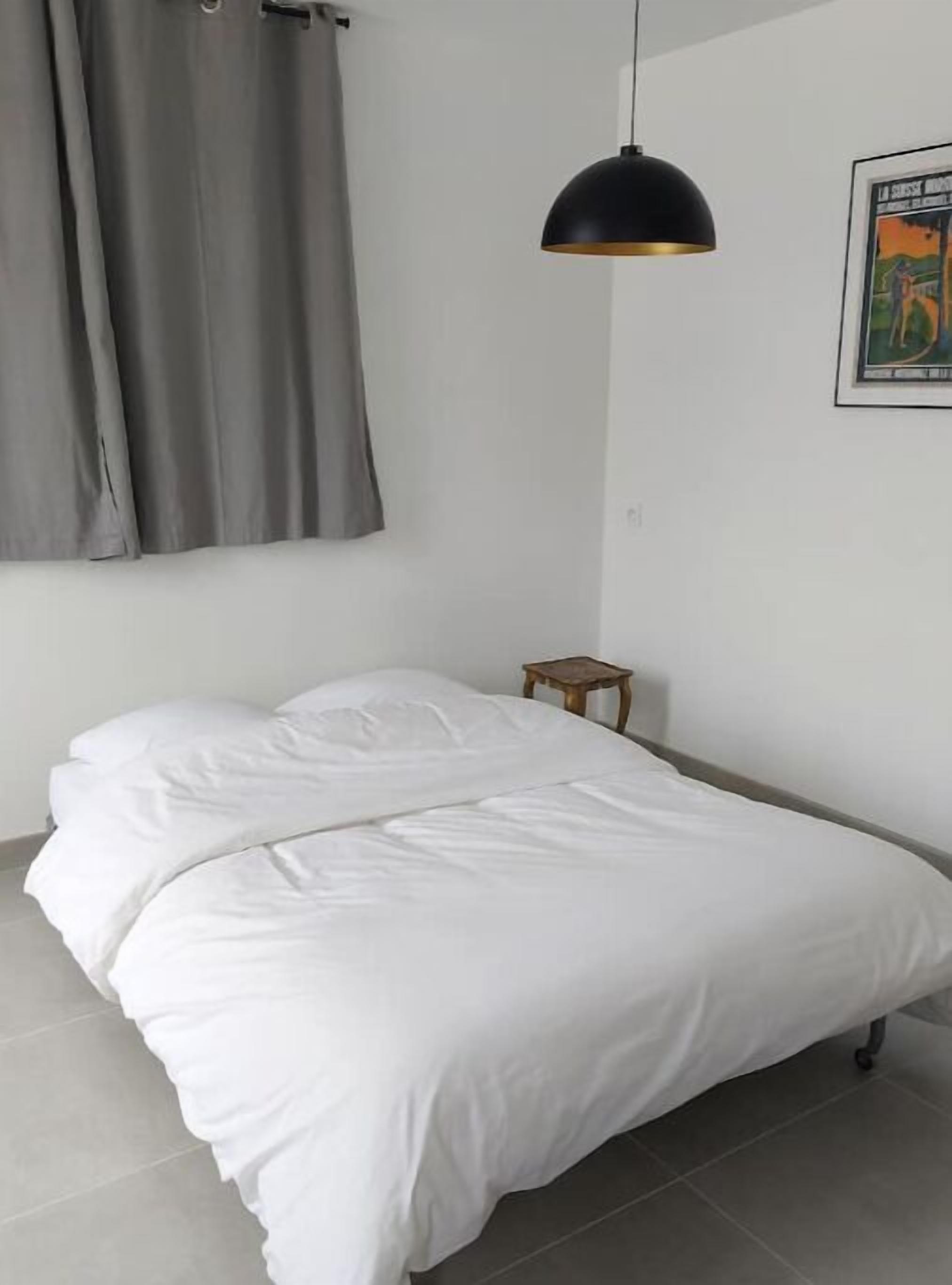 A bedroom with a bed, grey curtains, a small wooden nightstand, and a black pendant light.