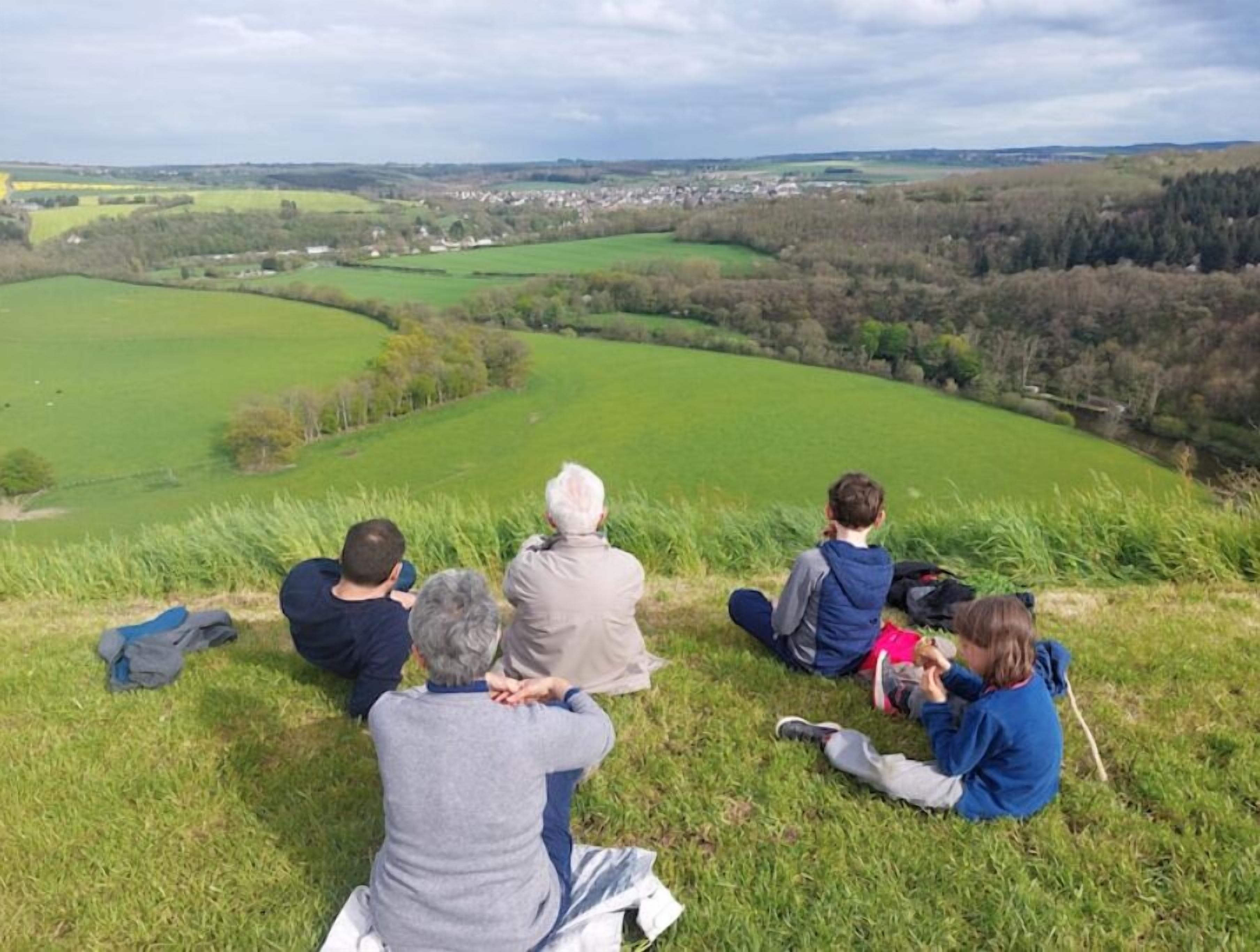 A group of people sitting on a grassy hill overlooking a rural landscape with fields and a small town in the distance.