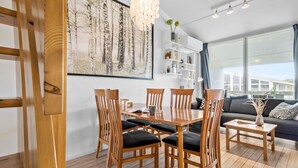 Een moderne woonkamer met een eethoek, met een groot raam, een houten eettafel met stoelen, een bank en een boekenkast.