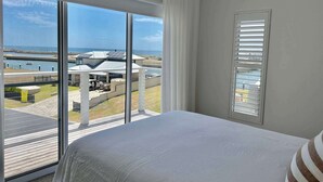 A bedroom with a large window, a bed, and a view of a coastal area with buildings and a body of water. - Casa Bianca - Ocean and harbour views (Cape Jaffa)