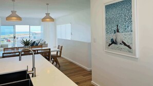 A modern kitchen with a white countertop, stainless steel sink, and wooden chairs. There is a dining area with a white table and chairs, and a large window offering a view of the ocean. - Casa Bianca - Ocean and harbour views (Cape Jaffa)