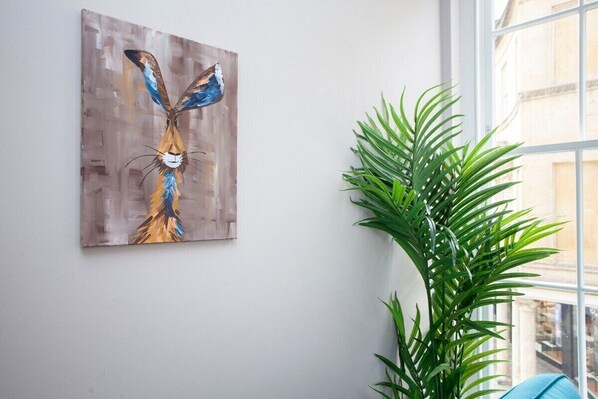 Pintura de un ave en la pared, una planta en maceta y una ventana con vistas a edificios.