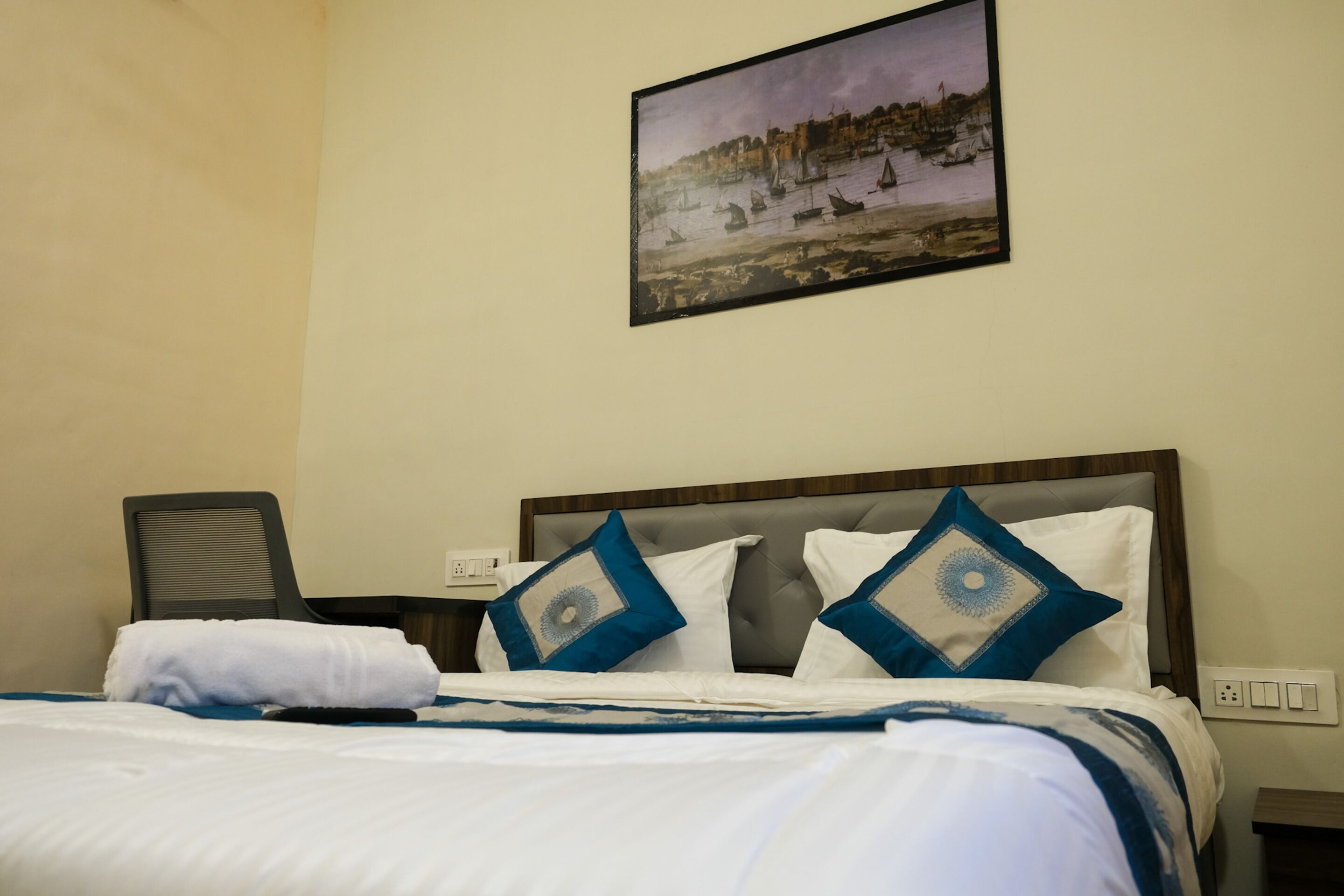 A hotel room with a bed, a painting of a harbor scene, and a chair.