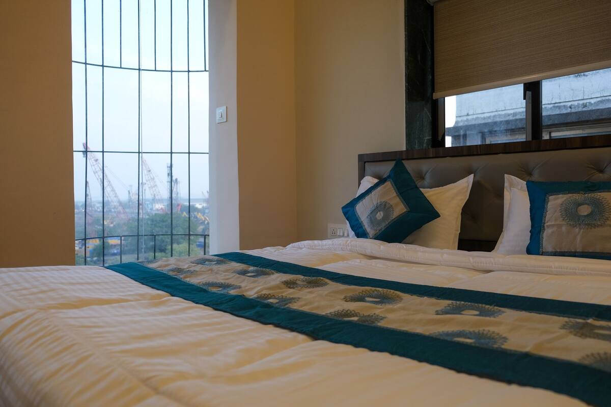 A hotel room with a large bed, a window with a view of construction cranes, and a headboard with decorative pillows.