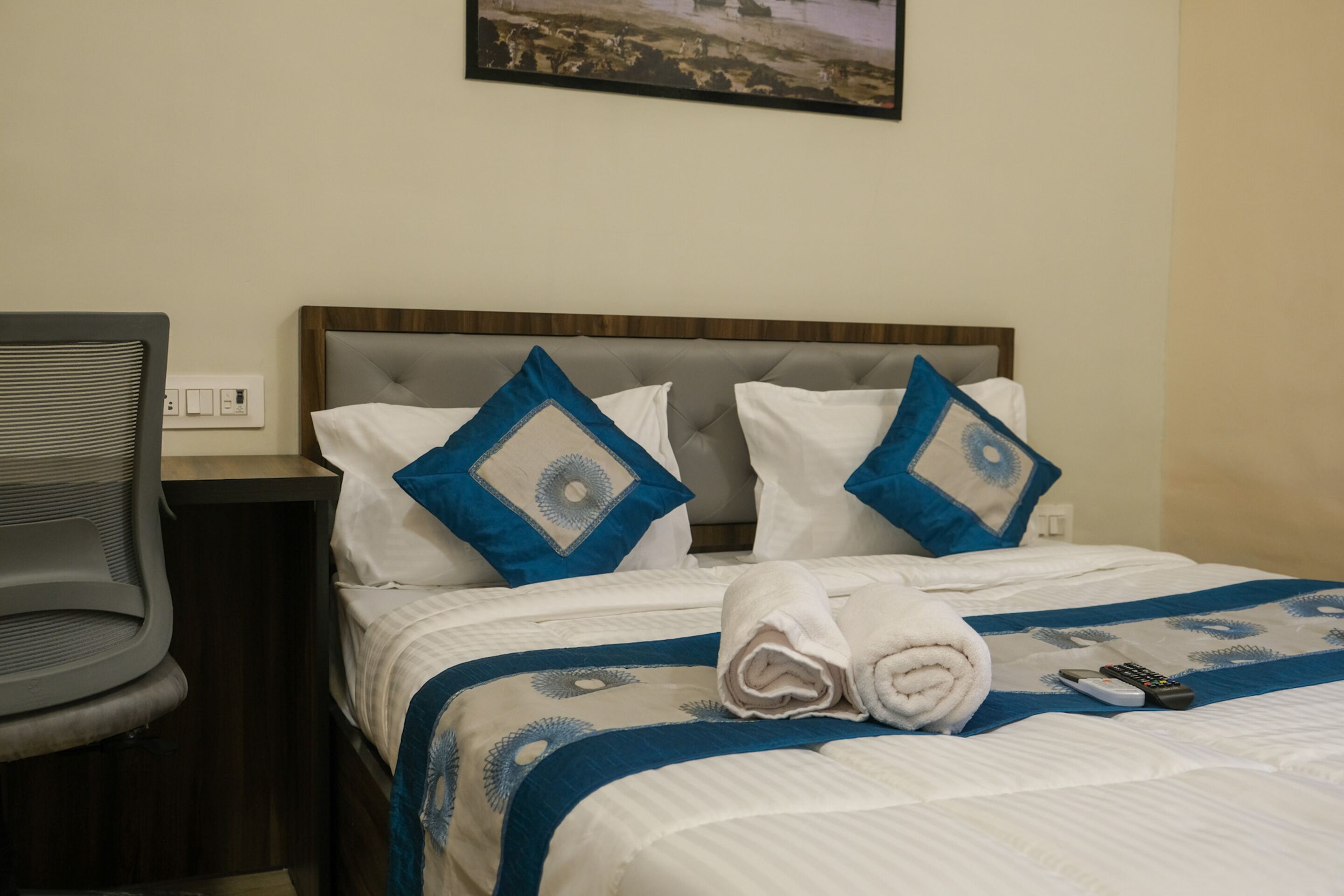 Una cama bien tendida con un edredón azul y blanco, dos almohadas con diseños azules y blancos, una toalla blanca enrollada y un control remoto sobre la cama.