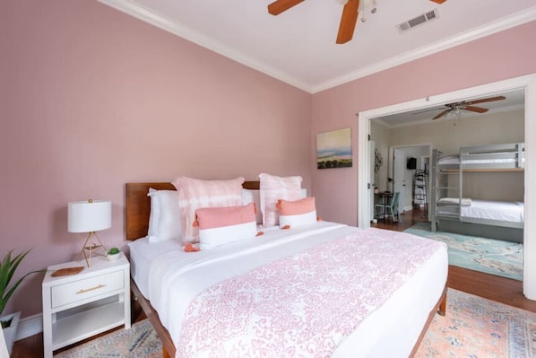 A bedroom with a bed, bedside table, and a ceiling fan. There is a door leading to another room with bunk beds.