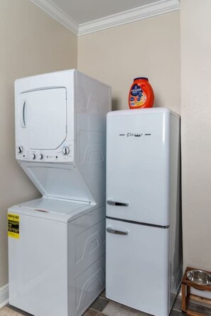 Une petite buanderie équipée d’un lave-linge à chargement frontal et d’un sèche-linge à chargement par le haut, d’un petit réfrigérateur et d’un saladier en métal posé sur un tabouret en bois.