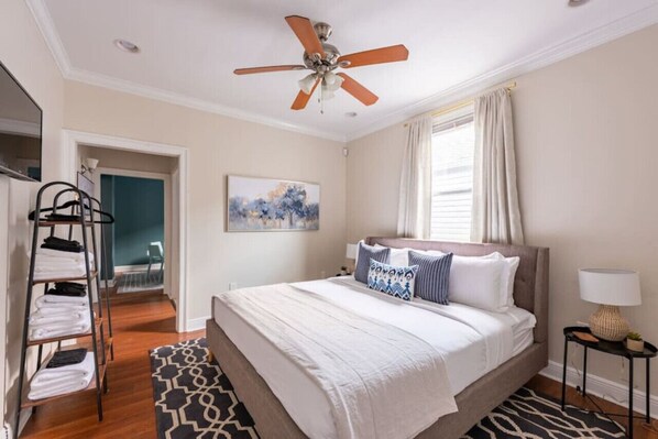 Une chambre à coucher comprenant un lit, un ventilateur de plafond, un tableau, une fenêtre avec des rideaux, une table de chevet avec une lampe et un porte-serviettes.