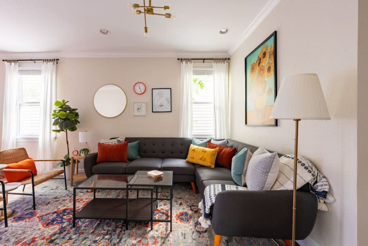 A modern living room with a sectional sofa, a coffee table, and a large rug. There is a plant, a lamp, and artwork on the walls.