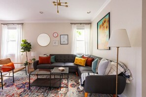Un salon moderne avec un canapé d’angle, une table basse et un grand tapis. On y trouve également une plante, une lampe et des œuvres d’art accrochées aux murs.